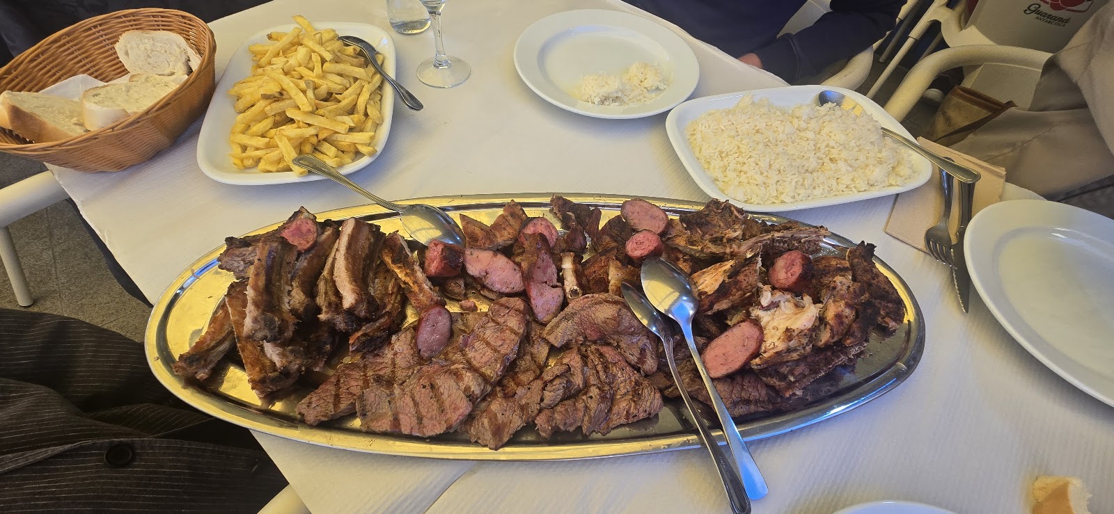 Travessa oval de grelhado misto com costeletas, chouriço e carnes fatiadas, servida com batata frita, arroz e pão.